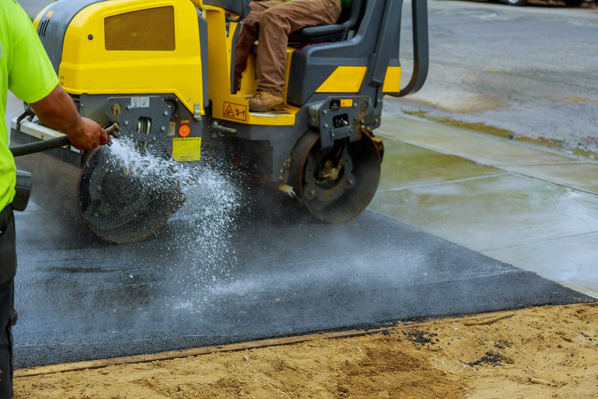 Residential paving crew using a roller machine and water spray to set fresh asphalt.